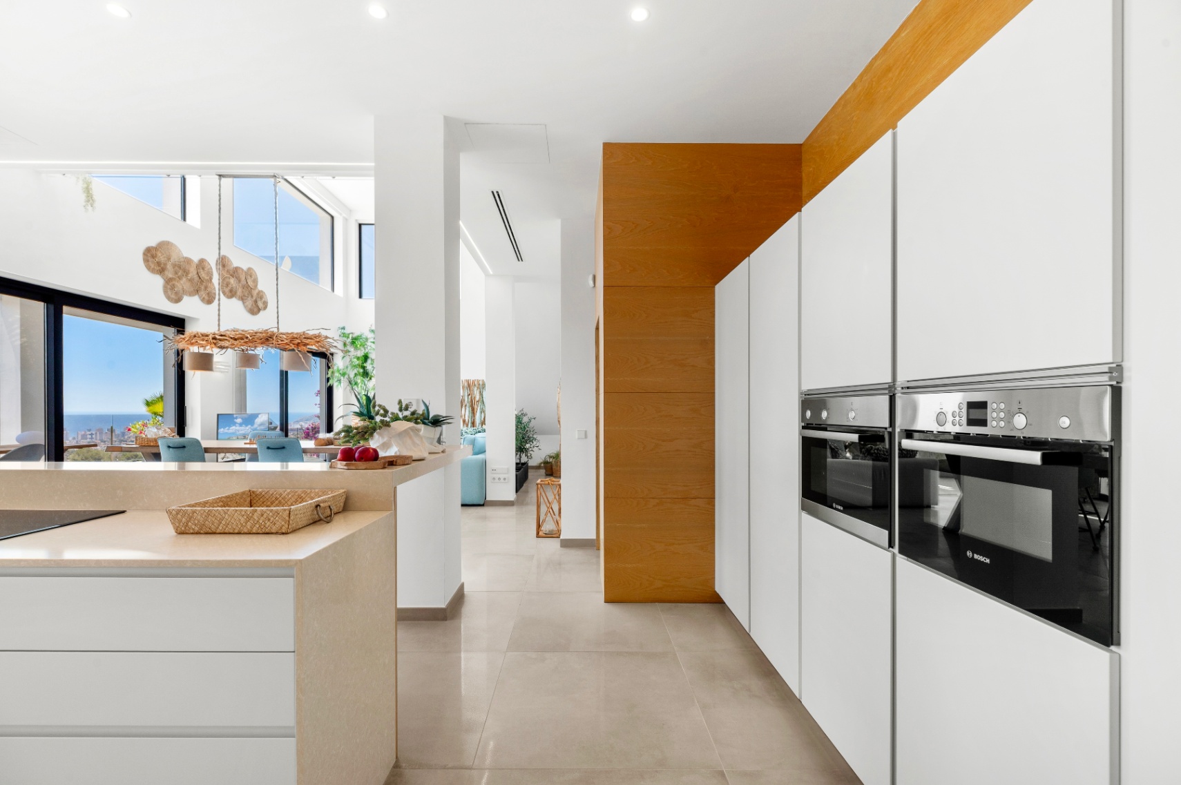 Elegante moderne Küche mit integrierten Bosch-Öfen, weißen grifflosen Schränken und heller Steininsel. Der offene Grundriss führt zu einem Essbereich mit Panoramameerblick durch große Glastüren. Elegante moderne Küche mit integrierten Bosch-Öfen, weißen grifflosen Schränken und heller Steininsel. Der offene Grundriss führt zu einem Essbereich mit Panoramameerblick durch große Glastüren.