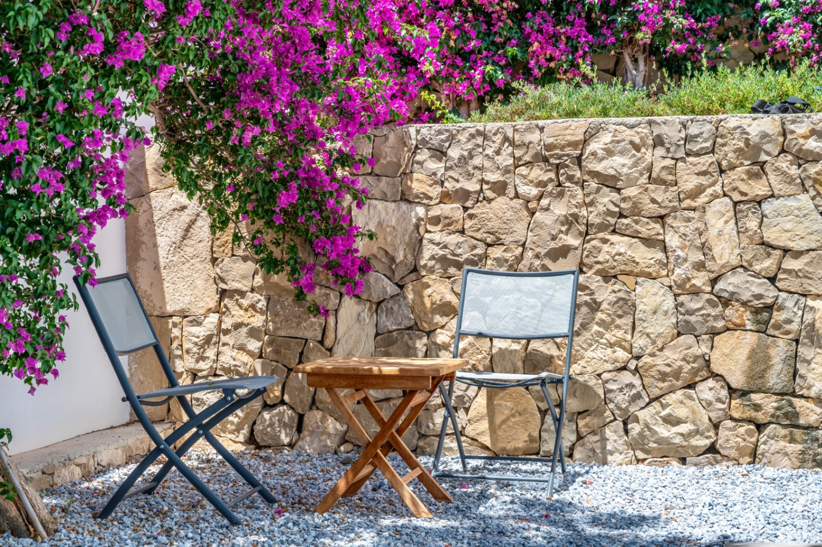 Gemütlicher Außenbereich mit zwei grauen Klappstühlen und Holztisch auf Kies. Eine Natursteinmauer wird von leuchtend magentafarbenen Bougainvilleen überrankt. Gemütlicher Außenbereich mit zwei grauen Klappstühlen und Holztisch auf Kies. Eine Natursteinmauer wird von leuchtend magentafarbenen Bougainvilleen überrankt.