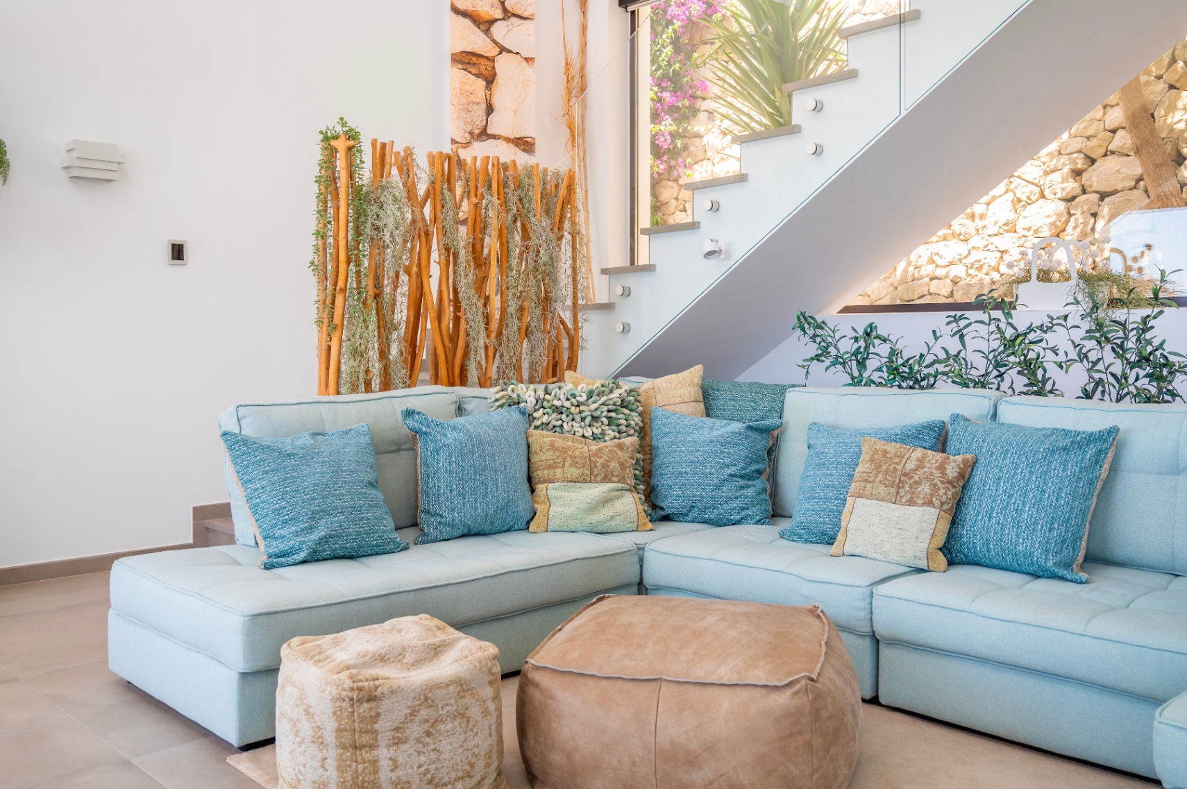 Gemütlicher Wohnbereich mit hellblauem Ecksofa, Kissen und zwei Lederpoufs. Moderne Glastreppe neben einem Naturholz-Pflanzen-Sichtschutz, mit Steinmauer im Hintergrund sichtbar. Gemütlicher Wohnbereich mit hellblauem Ecksofa, Kissen und zwei Lederpoufs. Moderne Glastreppe neben einem Naturholz-Pflanzen-Sichtschutz, mit Steinmauer im Hintergrund sichtbar.