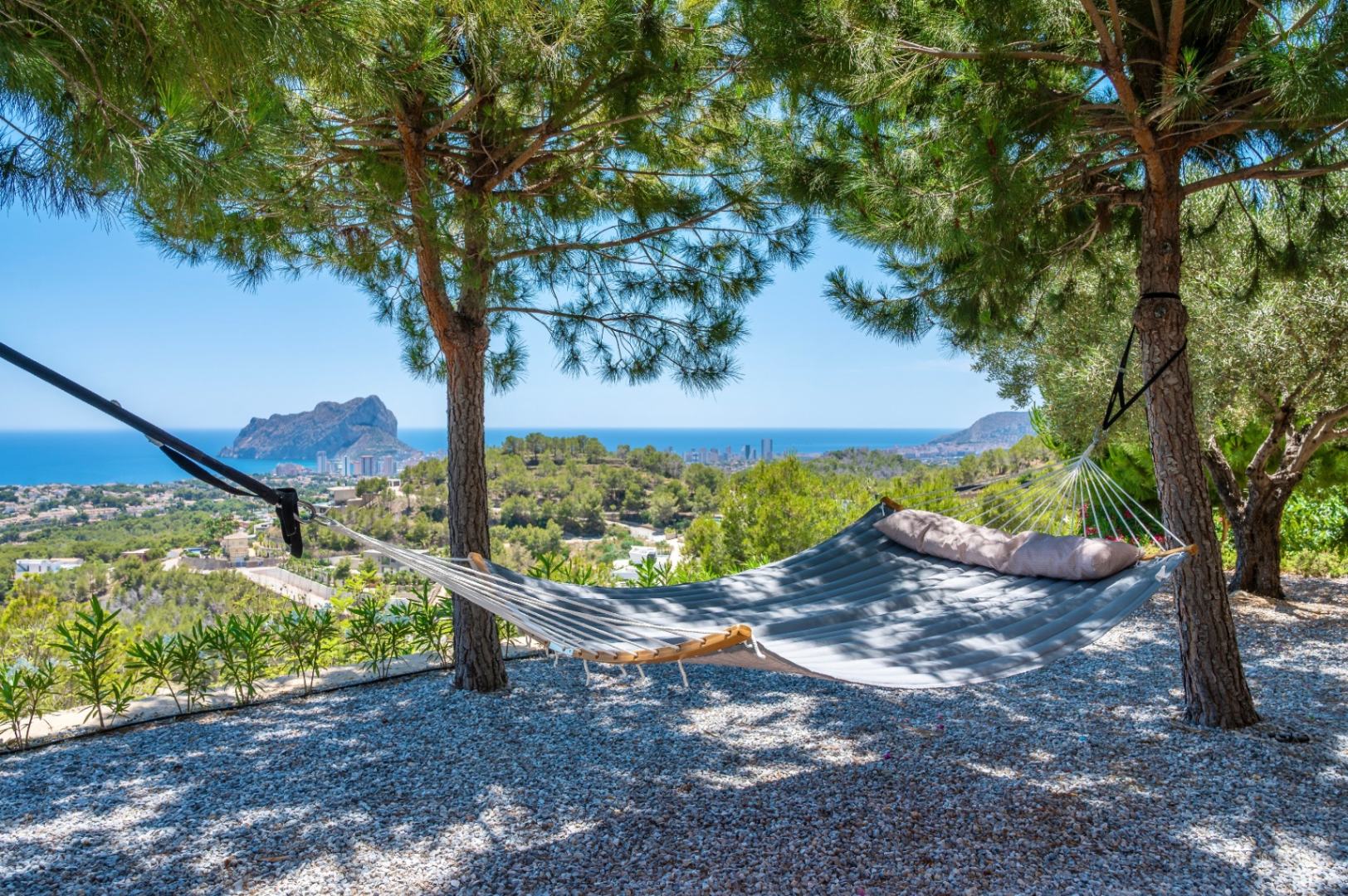 Gepolsterte Hängematte unter Kiefern, die einen ruhigen Rückzugsort mit Panoramablick auf das Mittelmeer und den markanten Peñon de Ifach bietet. Kiesboden. Gepolsterte Hängematte unter Kiefern, die einen ruhigen Rückzugsort mit Panoramablick auf das Mittelmeer und den markanten Peñon de Ifach bietet. Kiesboden.