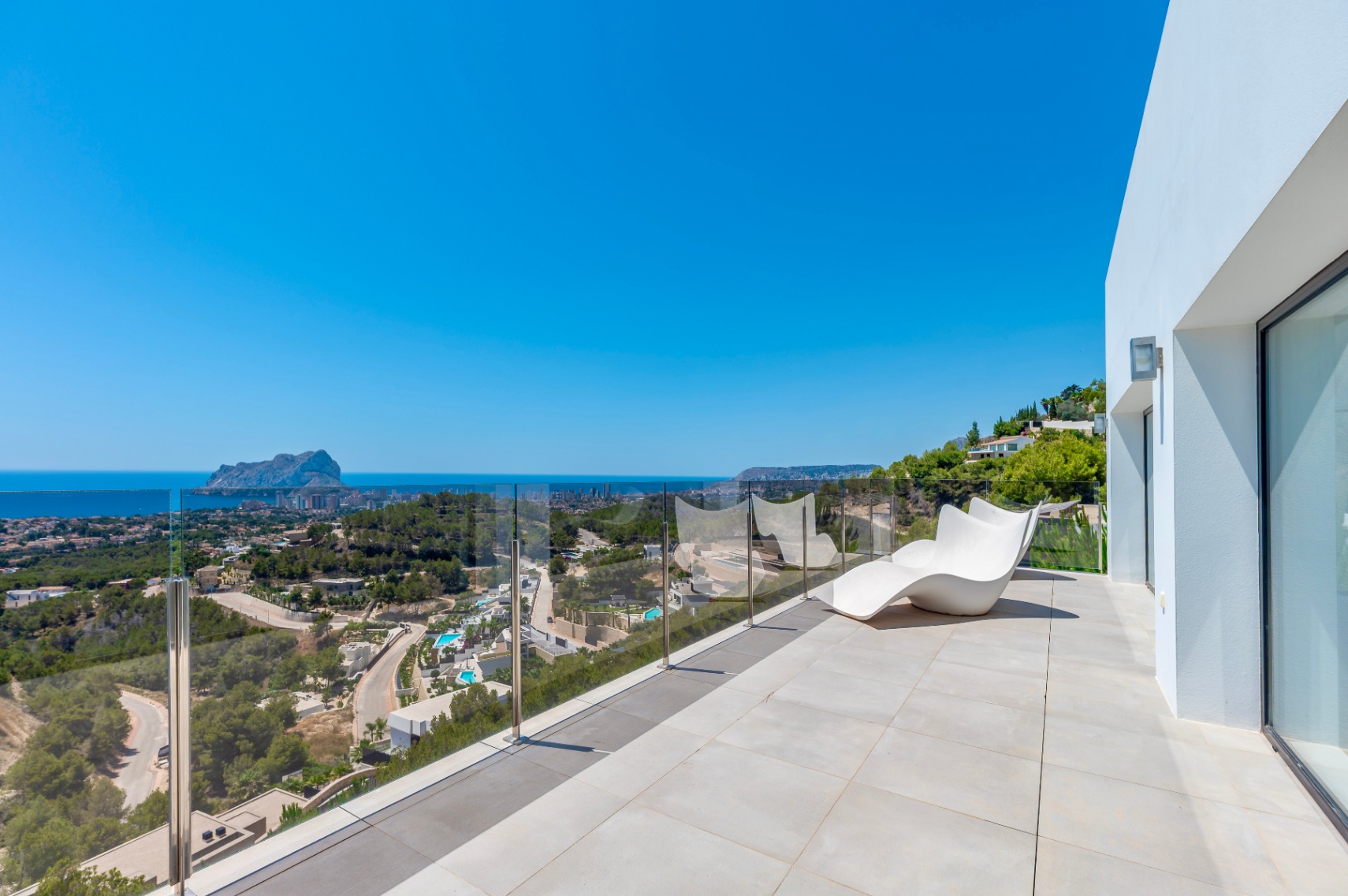Geräumige Außenterrasse mit hellen Fliesen, einem modernen weißen Liegestuhl und einer Glasbalustrade. Panoramablick auf das Mittelmeer, den Peñón de Ifach und die Küste von Calpe. Geräumige Außenterrasse mit hellen Fliesen, einem modernen weißen Liegestuhl und einer Glasbalustrade. Panoramablick auf das Mittelmeer, den Peñón de Ifach und die Küste von Calpe.