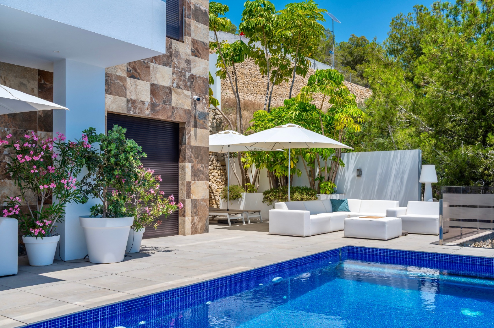 Geräumige Terrasse mit blauem Mosaikpool und weißen Gartenmöbeln unter Sonnenschirmen, ergänzt durch eine Steinfassade und üppige mediterrane Bepflanzung. Geräumige Terrasse mit blauem Mosaikpool und weißen Gartenmöbeln unter Sonnenschirmen, ergänzt durch eine Steinfassade und üppige mediterrane Bepflanzung.