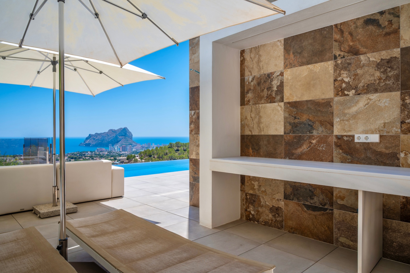 Großzügige Außenterrasse mit integrierter Theke, verziert mit Naturstein-Optikfliesen in Erdtönen. Panoramablick auf das Meer, den Infinity-Pool und den Peñon de Ifach. Großzügige Außenterrasse mit integrierter Theke, verziert mit Naturstein-Optikfliesen in Erdtönen. Panoramablick auf das Meer, den Infinity-Pool und den Peñon de Ifach.