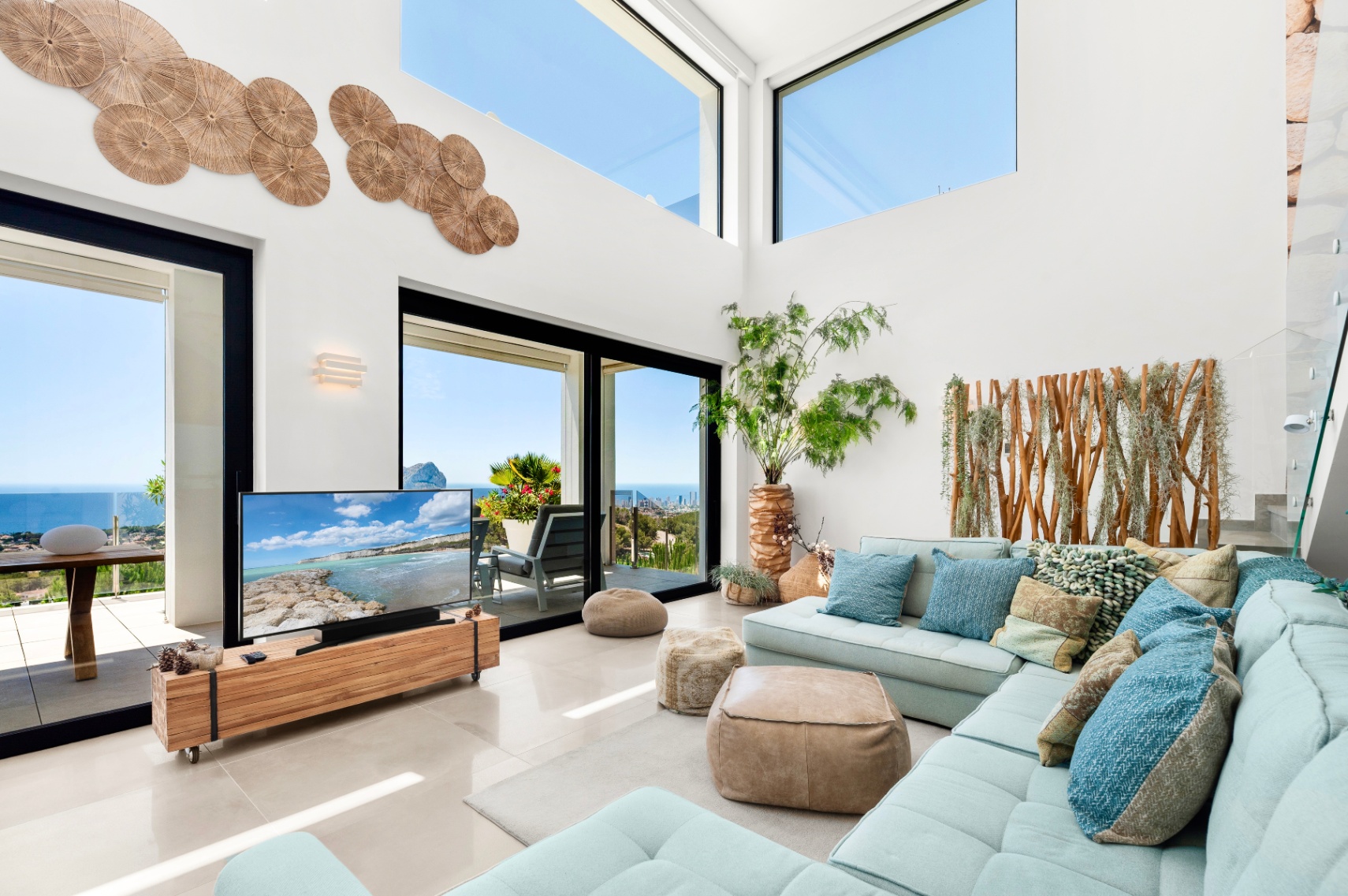 Großzügiges Wohnzimmer mit hohen Decken, großen Glasfronten und Oberlichtern, die einen weiten Meerblick ermöglichen. Ein hellblaues Ecksofa ergänzt Holzakzente und die individuelle Wandgestaltung. Großzügiges Wohnzimmer mit hohen Decken, großen Glasfronten und Oberlichtern, die einen weiten Meerblick ermöglichen. Ein hellblaues Ecksofa ergänzt Holzakzente und die individuelle Wandgestaltung.