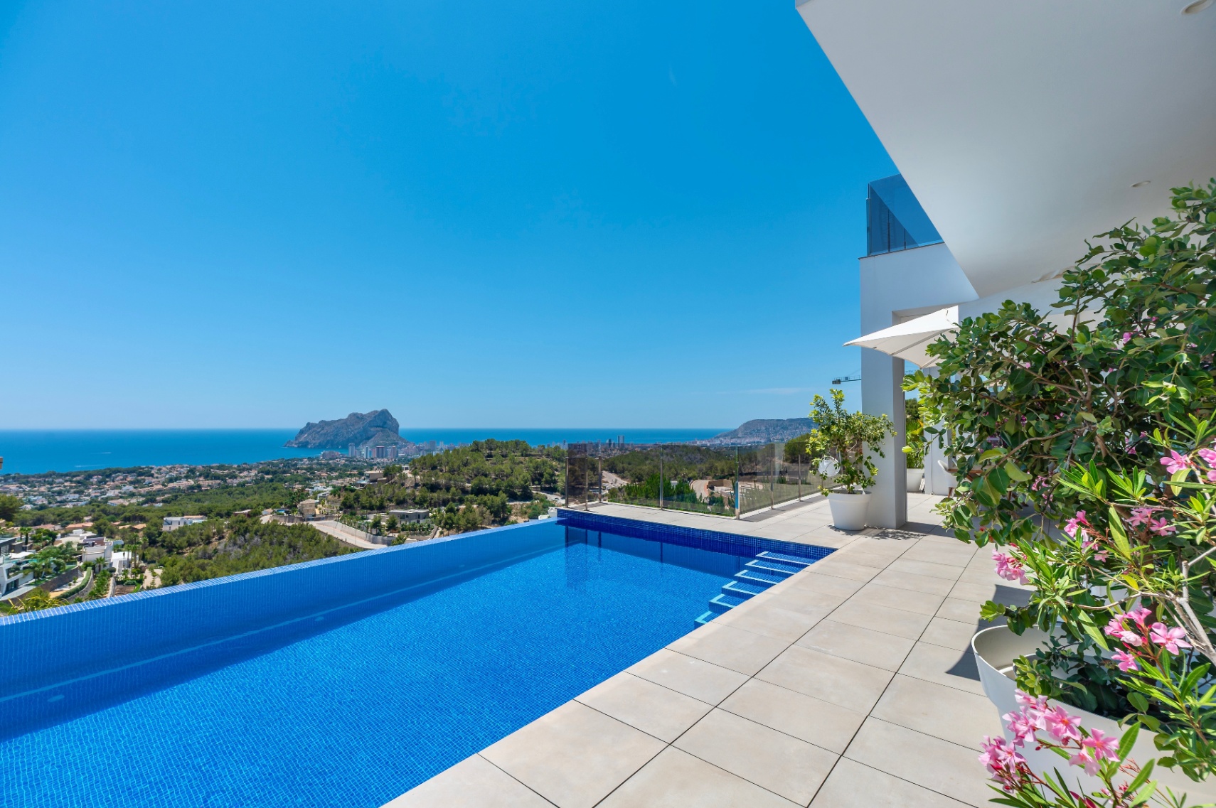 Infinity-Pool mit blauen Mosaikfliesen und heller Fliesenterrasse. Bietet freien Panoramablick auf das Meer und den Peñon de Ifach, ergänzt durch moderne Architektur und mediterrane Pflanzen. Infinity-Pool mit blauen Mosaikfliesen und heller Fliesenterrasse. Bietet freien Panoramablick auf das Meer und den Peñon de Ifach, ergänzt durch moderne Architektur und mediterrane Pflanzen.