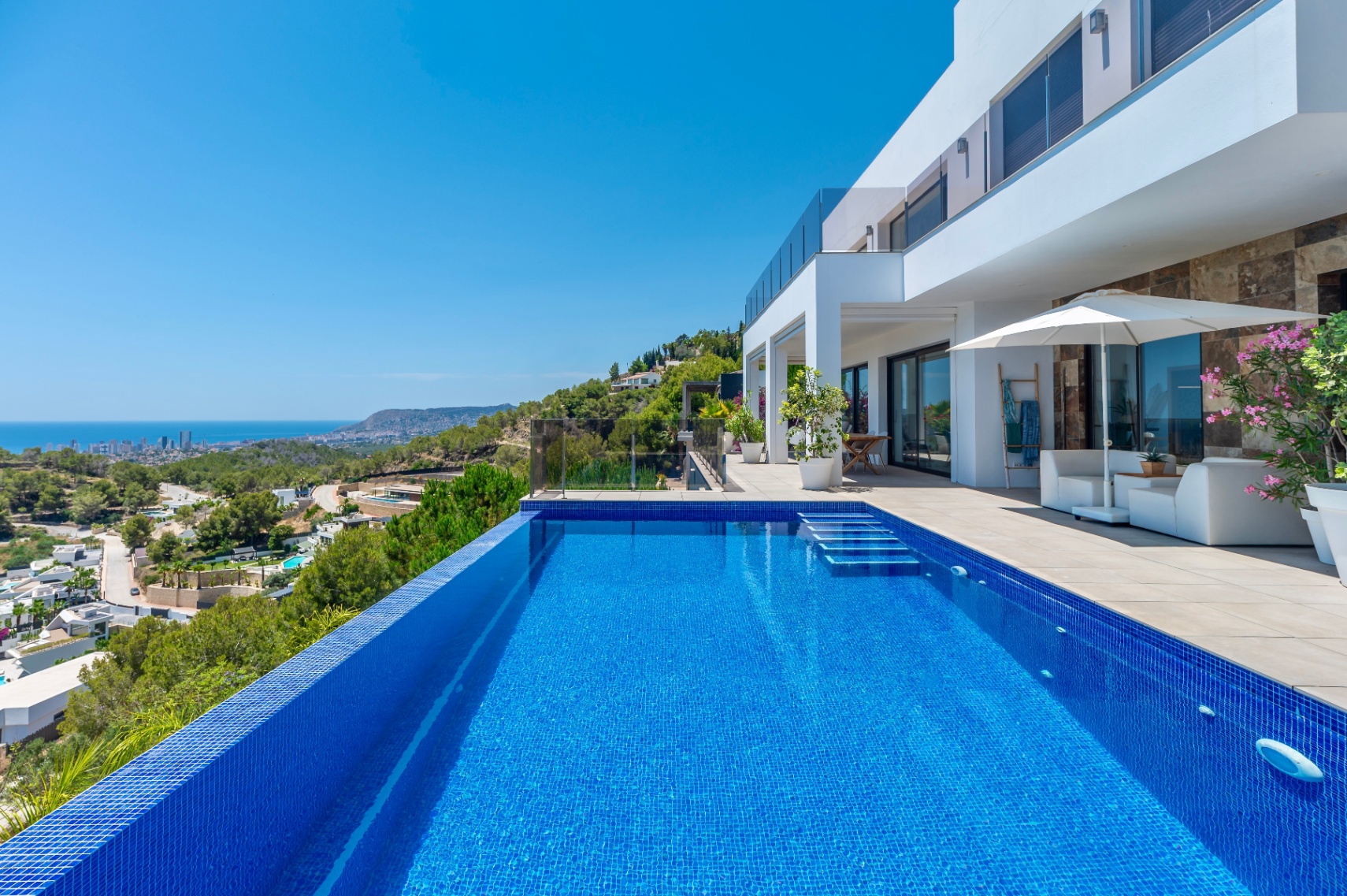 Infinity-Pool mit blauen Mosaikfliesen und integrierten Stufen, angrenzend an eine geräumige Terrasse mit weißer Lounge-Möblierung und Panoramablick auf Küste und Stadt. Infinity-Pool mit blauen Mosaikfliesen und integrierten Stufen, angrenzend an eine geräumige Terrasse mit weißer Lounge-Möblierung und Panoramablick auf Küste und Stadt.