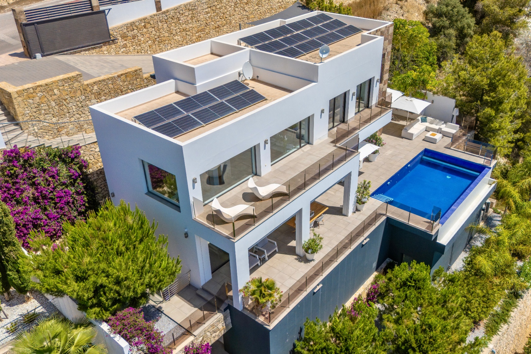 Luftaufnahme einer modernen weißen Villa mit mehreren Terrassenebenen, einem rechteckigen Pool und großzügigen Außenbereichen. Solarpaneele auf den Flachdächern, eingebettet in üppige mediterrane Vegetation. Luftaufnahme einer modernen weißen Villa mit mehreren Terrassenebenen, einem rechteckigen Pool und großzügigen Außenbereichen. Solarpaneele auf den Flachdächern, eingebettet in üppige mediterrane Vegetation.
