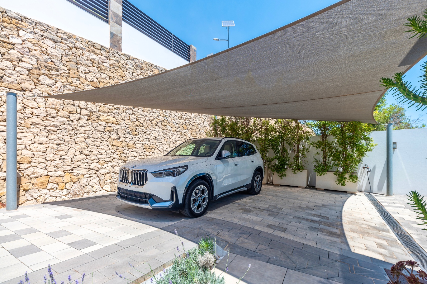 Moderner Außenparkplatz mit Sonnensegel über einer gepflasterten Einfahrt. Eine Natursteinmauer und begrünte Pflanzkübel umgeben den Stellplatz für ein weißes SUV. Moderner Außenparkplatz mit Sonnensegel über einer gepflasterten Einfahrt. Eine Natursteinmauer und begrünte Pflanzkübel umgeben den Stellplatz für ein weißes SUV.