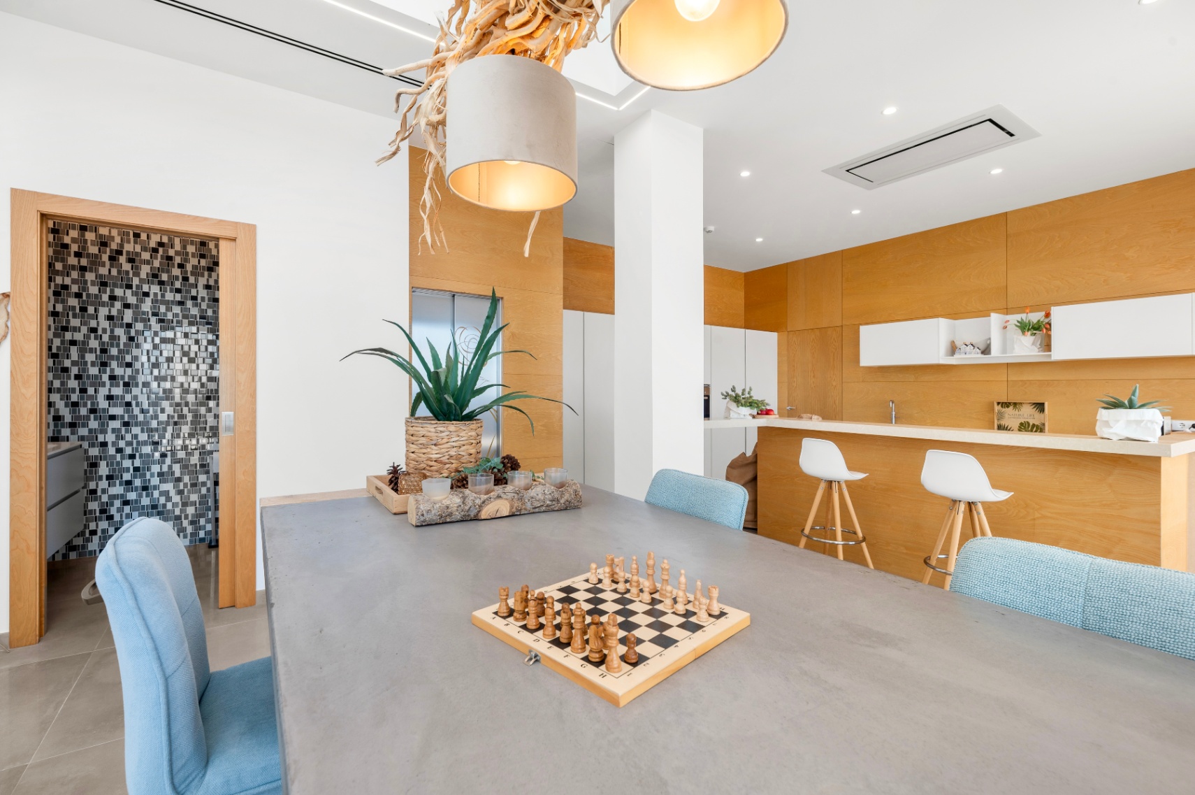 Moderner Essbereich mit Betontisch und hellblauen Stühlen. Eine markante Deckenleuchte aus Naturholz und eine offene Küche mit heller Holzverkleidung sind zu sehen. Zugang zu einem Mosaik-Badezimmer. Moderner Essbereich mit Betontisch und hellblauen Stühlen. Eine markante Deckenleuchte aus Naturholz und eine offene Küche mit heller Holzverkleidung sind zu sehen. Zugang zu einem Mosaik-Badezimmer.
