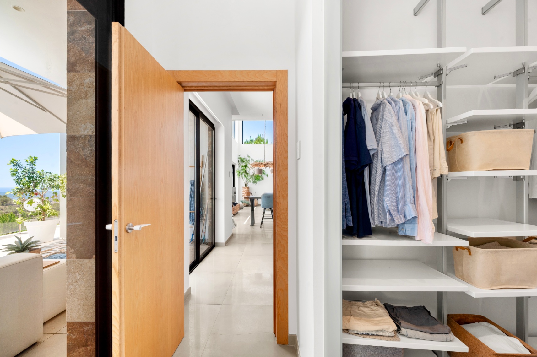 Moderner Flureingang mit offener heller Holztür, der einen weißen Einbauschrank mit verstellbaren Regalen und einen sonnigen Terrassenbereich mit Meerblick zeigt. Moderner Flureingang mit offener heller Holztür, der einen weißen Einbauschrank mit verstellbaren Regalen und einen sonnigen Terrassenbereich mit Meerblick zeigt.
