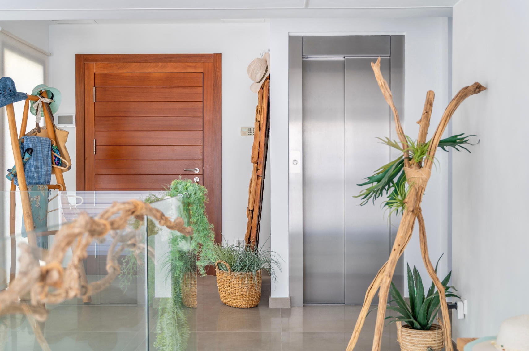 Moderner Innenflur mit eleganter Holztür und Edelstahlaufzug. Der Raum ist stilvoll mit natürlichen Elementen wie Treibholz, Topfpflanzen in Korbschalen und einer Glasbrüstung dekoriert. Moderner Innenflur mit eleganter Holztür und Edelstahlaufzug. Der Raum ist stilvoll mit natürlichen Elementen wie Treibholz, Topfpflanzen in Korbschalen und einer Glasbrüstung dekoriert.