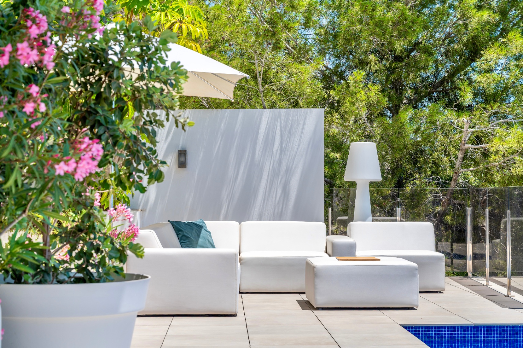 Moderner Outdoor-Loungebereich mit weißen Modulsofas und passendem Ottoman auf einer gefliesten Terrasse. Umgeben von üppigem Grün und einer Glasbrüstung neben dem Pool. Moderner Outdoor-Loungebereich mit weißen Modulsofas und passendem Ottoman auf einer gefliesten Terrasse. Umgeben von üppigem Grün und einer Glasbrüstung neben dem Pool.