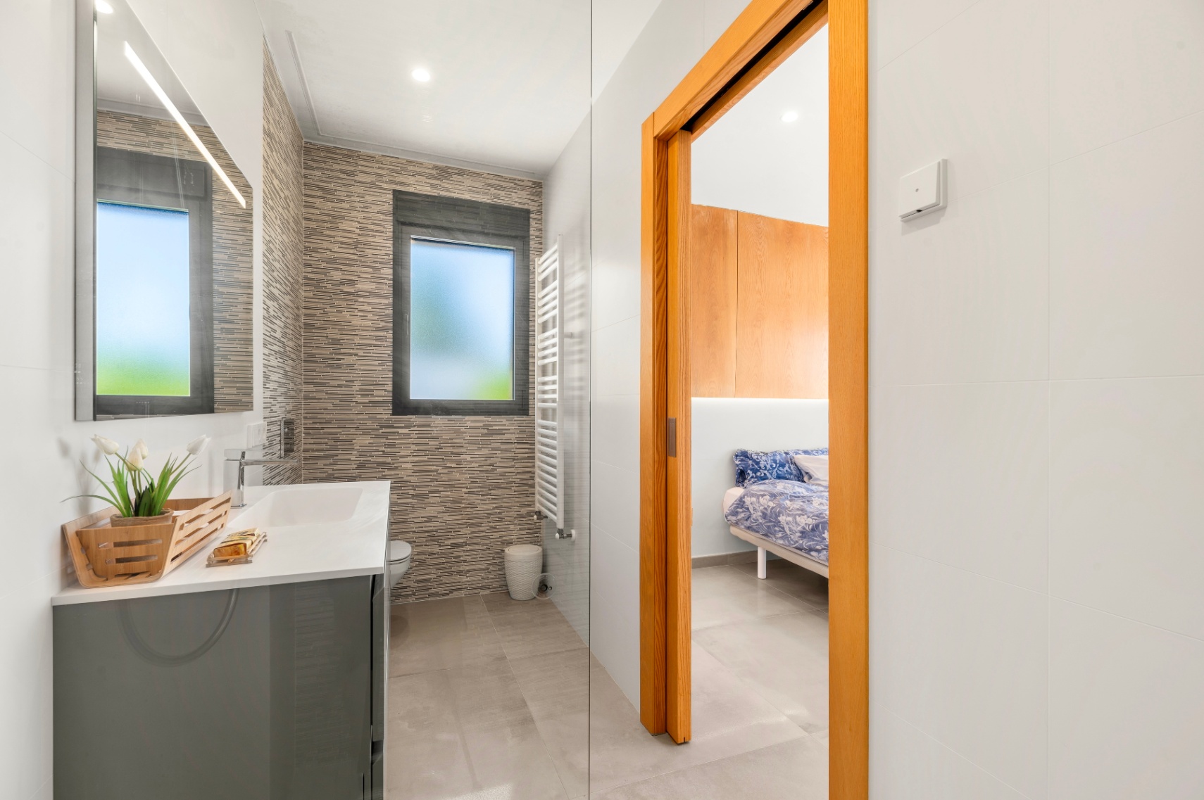 Modernes Badezimmer mit grauem Waschtisch, beleuchtetem Spiegel und strukturierter Wandverkleidung. Ein mattiertes Fenster und ein weißer Handtuchwärmer sind vorhanden, angrenzend an ein Schlafzimmer. Modernes Badezimmer mit grauem Waschtisch, beleuchtetem Spiegel und strukturierter Wandverkleidung. Ein mattiertes Fenster und ein weißer Handtuchwärmer sind vorhanden, angrenzend an ein Schlafzimmer.