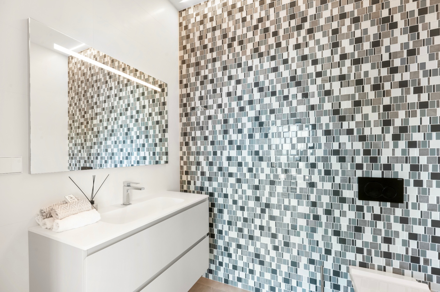 Modernes Badezimmer mit schwebendem weißen Waschtisch und integriertem Waschbecken. Die Akzentwand ist mit grau-weißen Mosaikfliesen verkleidet, ergänzt durch einen beleuchteten Spiegel. Modernes Badezimmer mit schwebendem weißen Waschtisch und integriertem Waschbecken. Die Akzentwand ist mit grau-weißen Mosaikfliesen verkleidet, ergänzt durch einen beleuchteten Spiegel.