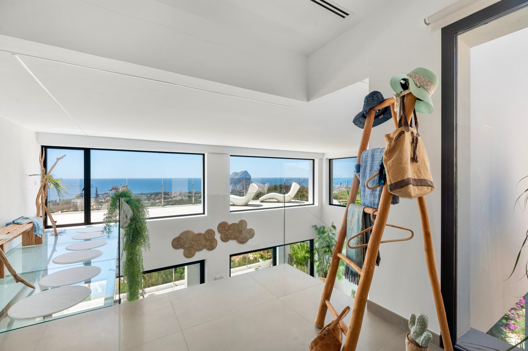 Obere Etage mit hellen Fliesen und Glasgeländer. Große Fenster bieten Panoramablicke auf Meer und Peñon de Ifach. Ein Glassteg mit ovalen Trittsteinen und eine dekorative Holzleiter sind sichtbar. Obere Etage mit hellen Fliesen und Glasgeländer. Große Fenster bieten Panoramablicke auf Meer und Peñon de Ifach. Ein Glassteg mit ovalen Trittsteinen und eine dekorative Holzleiter sind sichtbar.