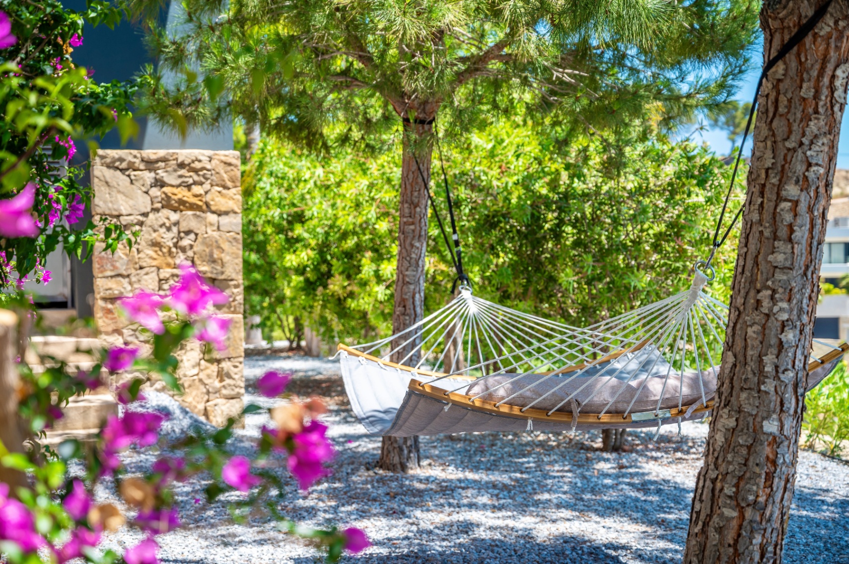 Ruhiger Gartenbereich mit einer bequemen grauen Hängematte mit Holzspreizstäben, zwischen Bäumen auf einem Kiesbett, umrahmt von lebhaften Bougainvilleen. Ruhiger Gartenbereich mit einer bequemen grauen Hängematte mit Holzspreizstäben, zwischen Bäumen auf einem Kiesbett, umrahmt von lebhaften Bougainvilleen.