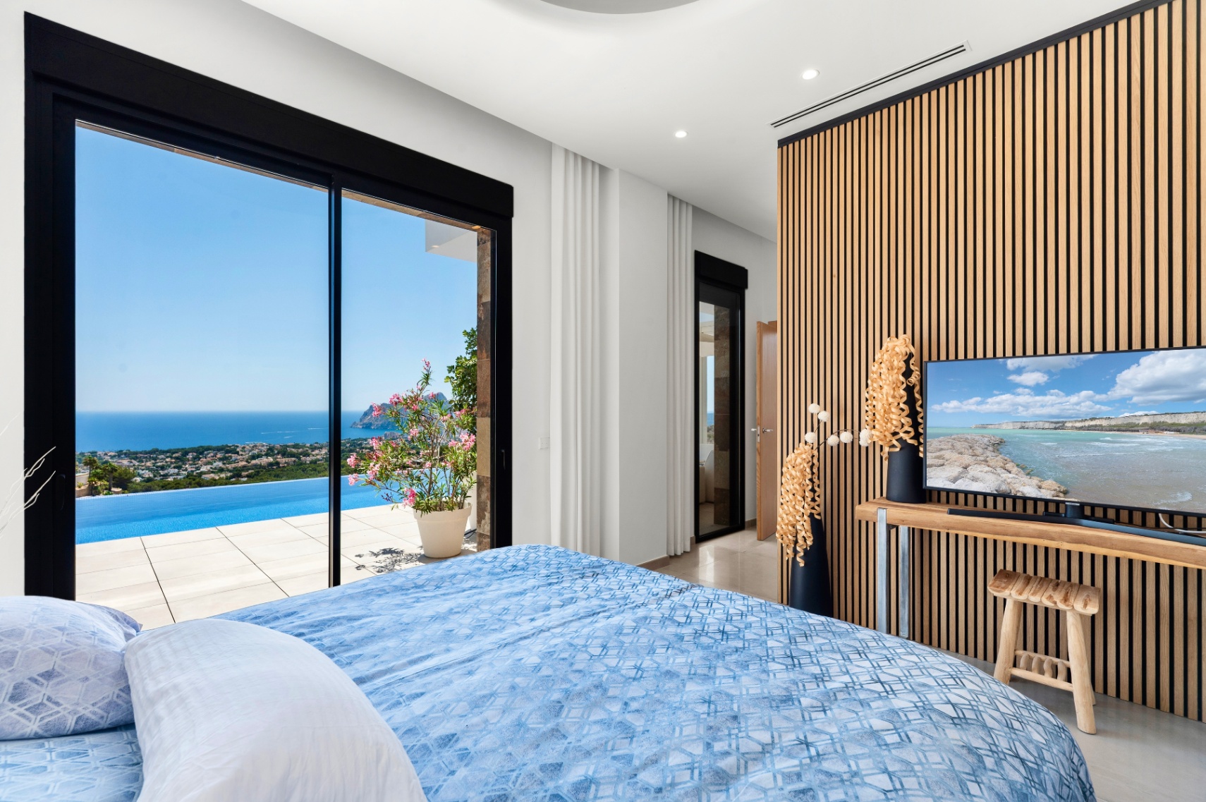 Schlafzimmer mit großen Glasschiebetüren, die sich zu einem Infinity-Pool und Panoramameerblick öffnen. Eine moderne Lattenwand aus Holz bildet den Hintergrund für den Fernseher und ergänzt das helle Interieur. Schlafzimmer mit großen Glasschiebetüren, die sich zu einem Infinity-Pool und Panoramameerblick öffnen. Eine moderne Lattenwand aus Holz bildet den Hintergrund für den Fernseher und ergänzt das helle Interieur.