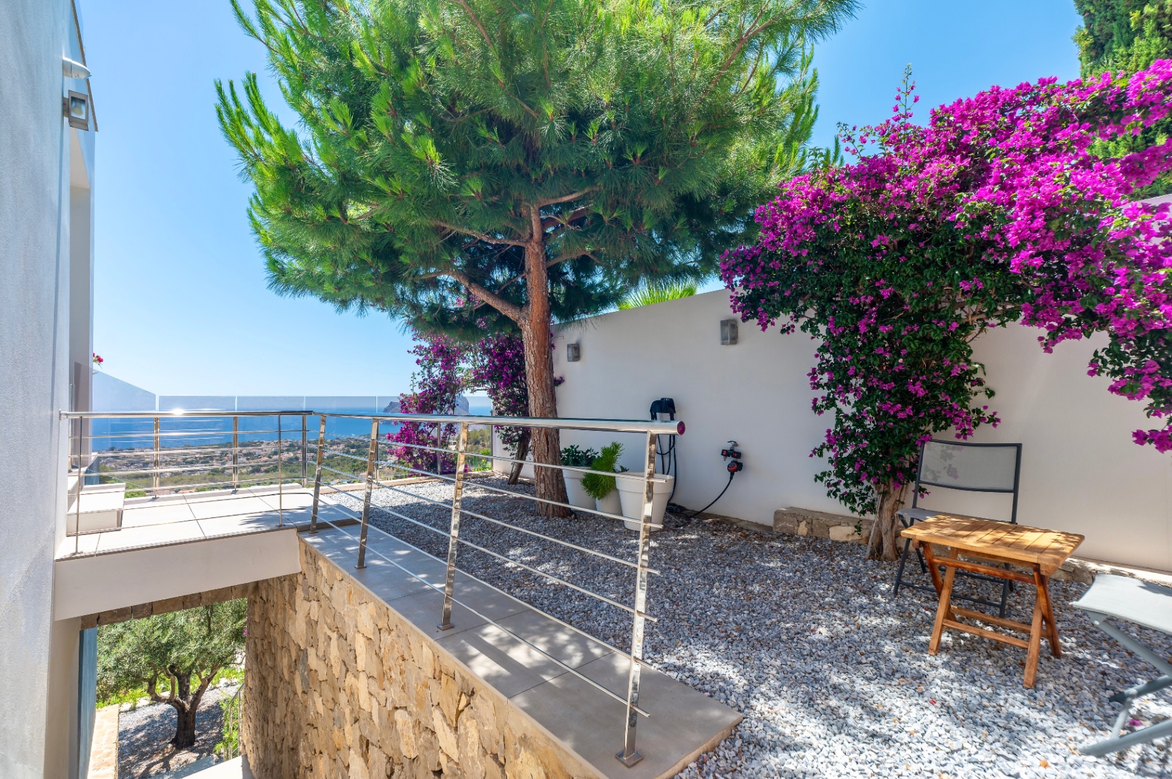 Terrasse mit moderner Edelstahl- und Glasbrüstung, bietet Panoramablick auf das Meer. Angrenzender Kiesgarten mit großer Kiefer und leuchtend lila Bougainvillea an einer weißen Wand. Terrasse mit moderner Edelstahl- und Glasbrüstung, bietet Panoramablick auf das Meer. Angrenzender Kiesgarten mit großer Kiefer und leuchtend lila Bougainvillea an einer weißen Wand.