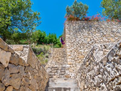 Außensteintreppe, umgeben von Trockenmauern aus Naturstein, die sich zwischen üppiger mediterraner Vegetation unter einem strahlend blauen Himmel emporwindet.