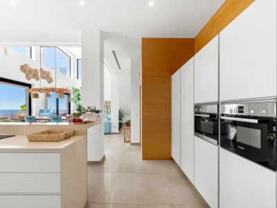 Elegante moderne Küche mit integrierten Bosch-Öfen, weißen grifflosen Schränken und heller Steininsel. Der offene Grundriss führt zu einem Essbereich mit Panoramameerblick durch große Glastüren.