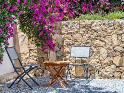 Gemütlicher Außenbereich mit zwei grauen Klappstühlen und Holztisch auf Kies. Eine Natursteinmauer wird von leuchtend magentafarbenen Bougainvilleen überrankt.