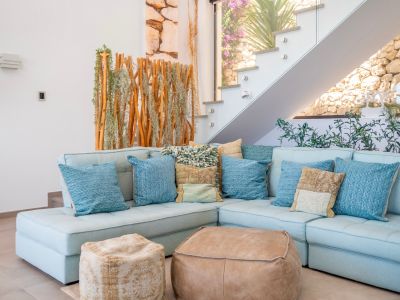Gemütlicher Wohnbereich mit hellblauem Ecksofa, Kissen und zwei Lederpoufs. Moderne Glastreppe neben einem Naturholz-Pflanzen-Sichtschutz, mit Steinmauer im Hintergrund sichtbar.