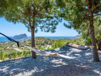 Gepolsterte Hängematte unter Kiefern, die einen ruhigen Rückzugsort mit Panoramablick auf das Mittelmeer und den markanten Peñon de Ifach bietet. Kiesboden.
