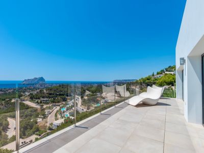 Geräumige Außenterrasse mit hellen Fliesen, einem modernen weißen Liegestuhl und einer Glasbalustrade. Panoramablick auf das Mittelmeer, den Peñón de Ifach und die Küste von Calpe.