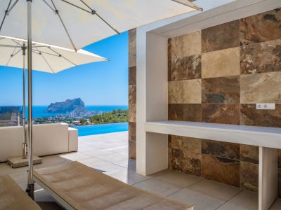 Großzügige Außenterrasse mit integrierter Theke, verziert mit Naturstein-Optikfliesen in Erdtönen. Panoramablick auf das Meer, den Infinity-Pool und den Peñon de Ifach.