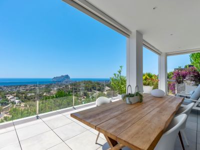 Großzügige überdachte Terrasse mit einem Massivholztisch und weißen Stühlen. Bietet Panoramablick auf das Meer, Calpe und den Peñón de Ifach durch eine Glasbrüstung.