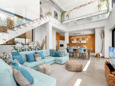 Großzügiger offener Wohnbereich mit hellblauem Ecksofa, moderner Glasgeländer-Treppe und Küche mit warmen Holzdetails, alles durchflutet von Tageslicht.