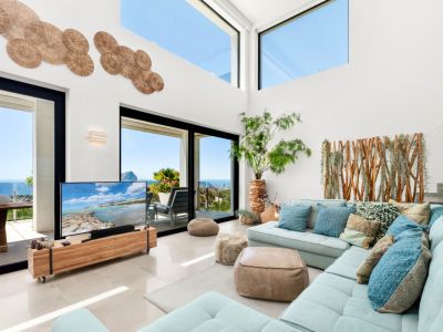Großzügiges Wohnzimmer mit hohen Decken, großen Glasfronten und Oberlichtern, die einen weiten Meerblick ermöglichen. Ein hellblaues Ecksofa ergänzt Holzakzente und die individuelle Wandgestaltung.