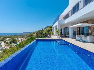 Infinity-Pool mit blauen Mosaikfliesen und integrierten Stufen, angrenzend an eine geräumige Terrasse mit weißer Lounge-Möblierung und Panoramablick auf Küste und Stadt.