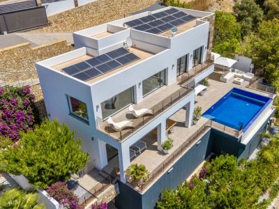 Luftaufnahme einer modernen weißen Villa mit mehreren Terrassenebenen, einem rechteckigen Pool und großzügigen Außenbereichen. Solarpaneele auf den Flachdächern, eingebettet in üppige mediterrane Vegetation.