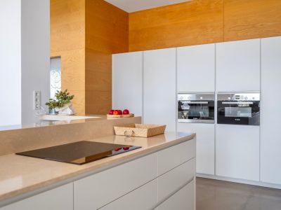 Moderne Küche mit sandfarbener Arbeitsplatte und integriertem Induktionskochfeld. Weiße grifflose Schränke und zwei eingebaute Bosch-Öfen sind sichtbar.