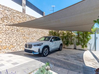 Moderner Außenparkplatz mit Sonnensegel über einer gepflasterten Einfahrt. Eine Natursteinmauer und begrünte Pflanzkübel umgeben den Stellplatz für ein weißes SUV.