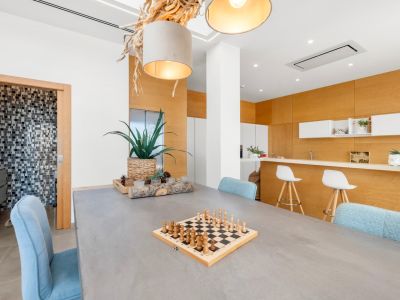 Moderner Essbereich mit Betontisch und hellblauen Stühlen. Eine markante Deckenleuchte aus Naturholz und eine offene Küche mit heller Holzverkleidung sind zu sehen. Zugang zu einem Mosaik-Badezimmer.