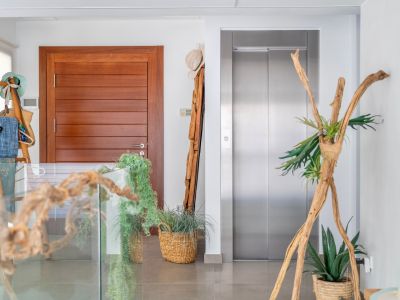 Moderner Innenflur mit eleganter Holztür und Edelstahlaufzug. Der Raum ist stilvoll mit natürlichen Elementen wie Treibholz, Topfpflanzen in Korbschalen und einer Glasbrüstung dekoriert.