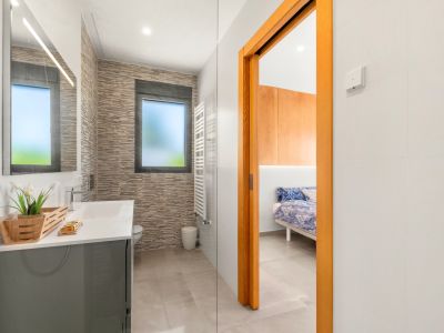 Modernes Badezimmer mit grauem Waschtisch, beleuchtetem Spiegel und strukturierter Wandverkleidung. Ein mattiertes Fenster und ein weißer Handtuchwärmer sind vorhanden, angrenzend an ein Schlafzimmer.