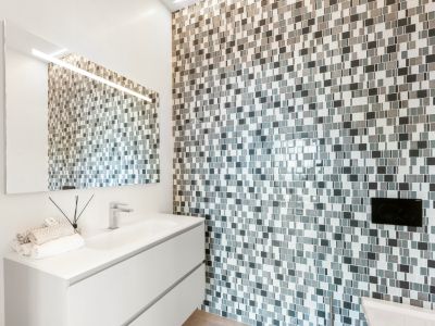 Modernes Badezimmer mit schwebendem weißen Waschtisch und integriertem Waschbecken. Die Akzentwand ist mit grau-weißen Mosaikfliesen verkleidet, ergänzt durch einen beleuchteten Spiegel.