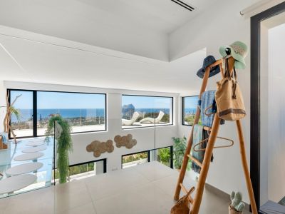 Obere Etage mit hellen Fliesen und Glasgeländer. Große Fenster bieten Panoramablicke auf Meer und Peñon de Ifach. Ein Glassteg mit ovalen Trittsteinen und eine dekorative Holzleiter sind sichtbar.