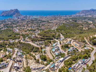 Panoramische Luftaufnahme von Calpe, mit dem Peñon de Ifach, der Küste und einem Wohngebiet mit Villen und Pools in grünen Hügeln und Serpentinenstraßen.