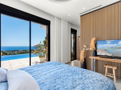 Schlafzimmer mit großen Glasschiebetüren, die sich zu einem Infinity-Pool und Panoramameerblick öffnen. Eine moderne Lattenwand aus Holz bildet den Hintergrund für den Fernseher und ergänzt das helle Interieur.