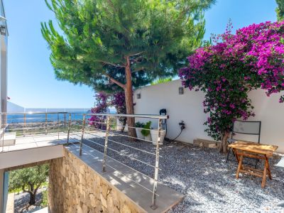 Terrasse mit moderner Edelstahl- und Glasbrüstung, bietet Panoramablick auf das Meer. Angrenzender Kiesgarten mit großer Kiefer und leuchtend lila Bougainvillea an einer weißen Wand.
