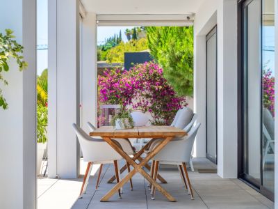 Überdachte Außenterrasse mit Holzesstisch und weißen Stühlen, umgeben von üppigem Grün und leuchtenden Bougainvillea, perfekt für Mahlzeiten im Freien.