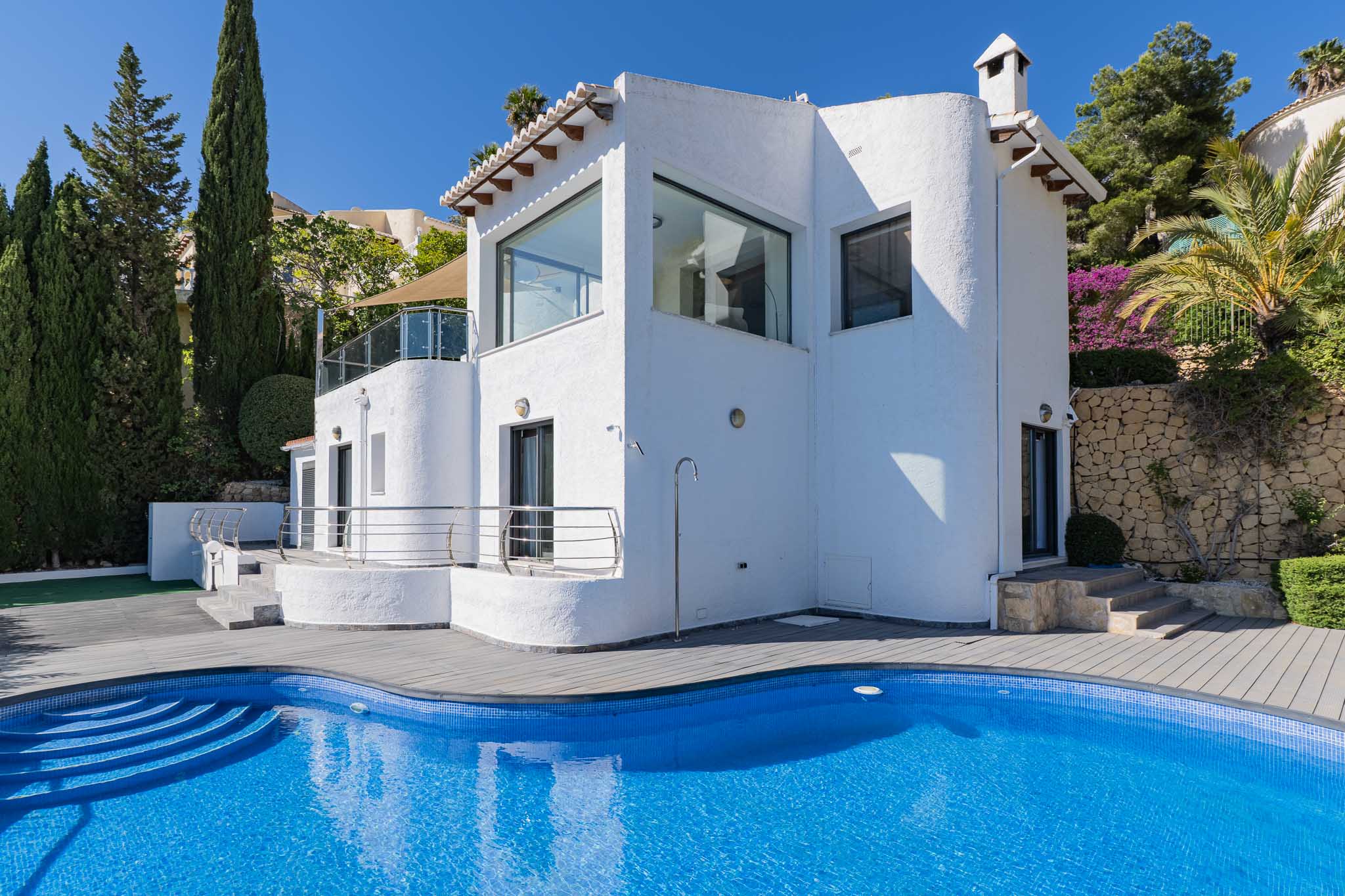 Außenansicht der Villa mit geschwungenem blauen Mosaikpool, grauer Verbundterrasse, Außendusche und mediterraner Bepflanzung.