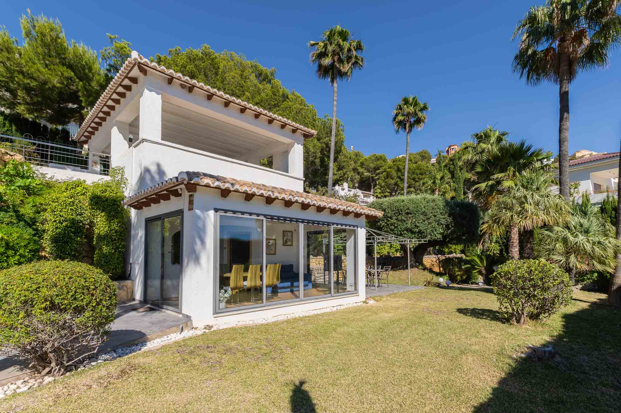 Außenansicht der Villa: weiße Fassade, traditionelle Ziegel, große Glasfronten. Der flache Garten bietet Rasenfläche und eine Terrasse mit Pergola, umgeben von Palmen und Büschen.