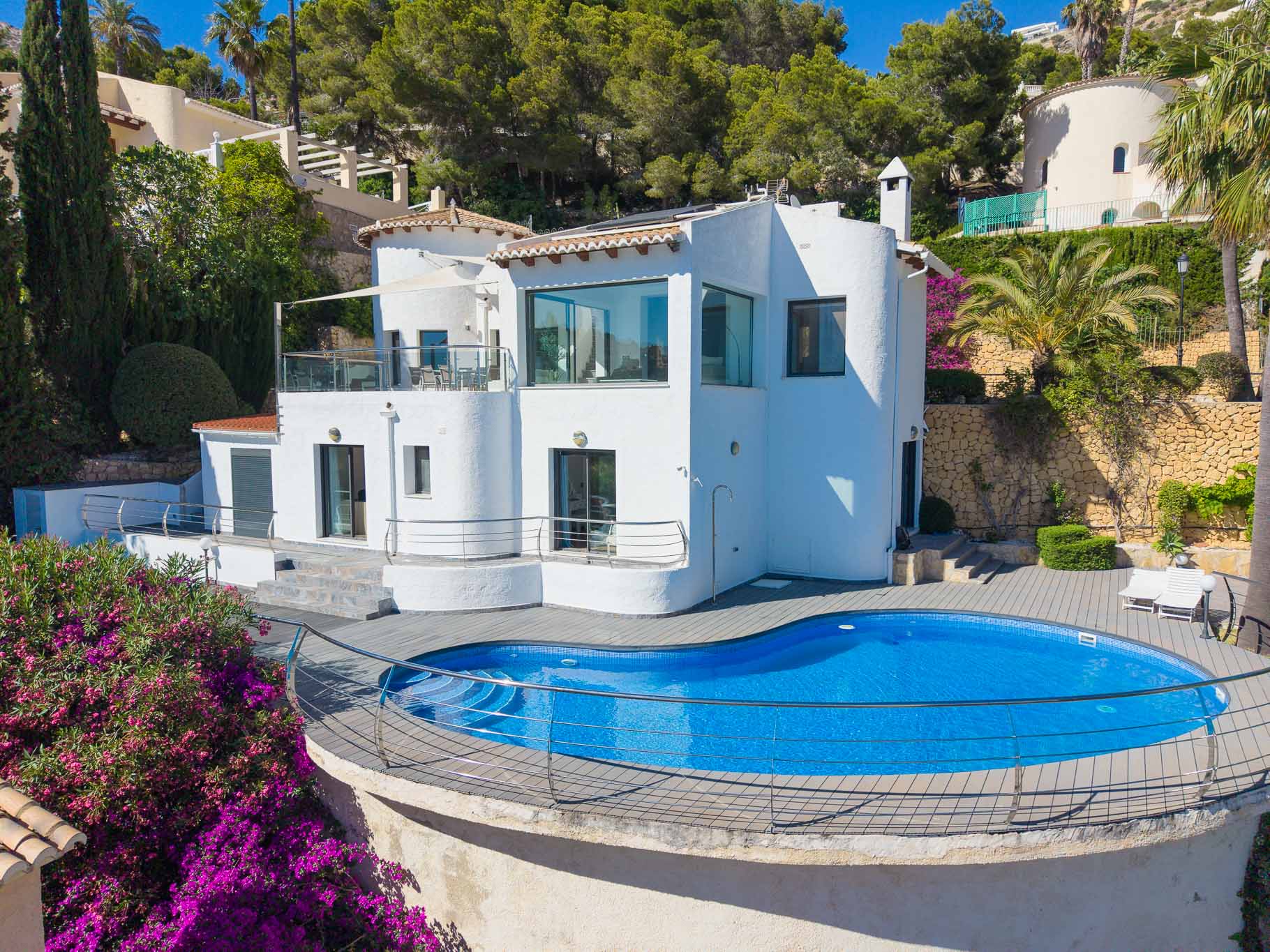 Außenansicht der weißen Villa mit geschwungenem Pool und Holzdeck. Die obere Terrasse mit Sonnenschutz und leuchtende Bougainvillea prägen das mediterrane Ambiente.
