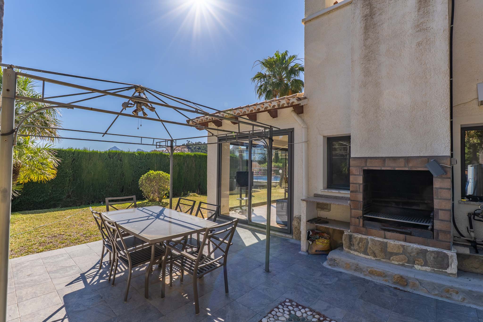Außenterrasse mit dunkelgrauen Fliesen, Metallpergola und Essgruppe. Ein gemauerter Grill aus Ziegeln und Stein sowie Glasschiebetüren zum Innenbereich sind sichtbar.