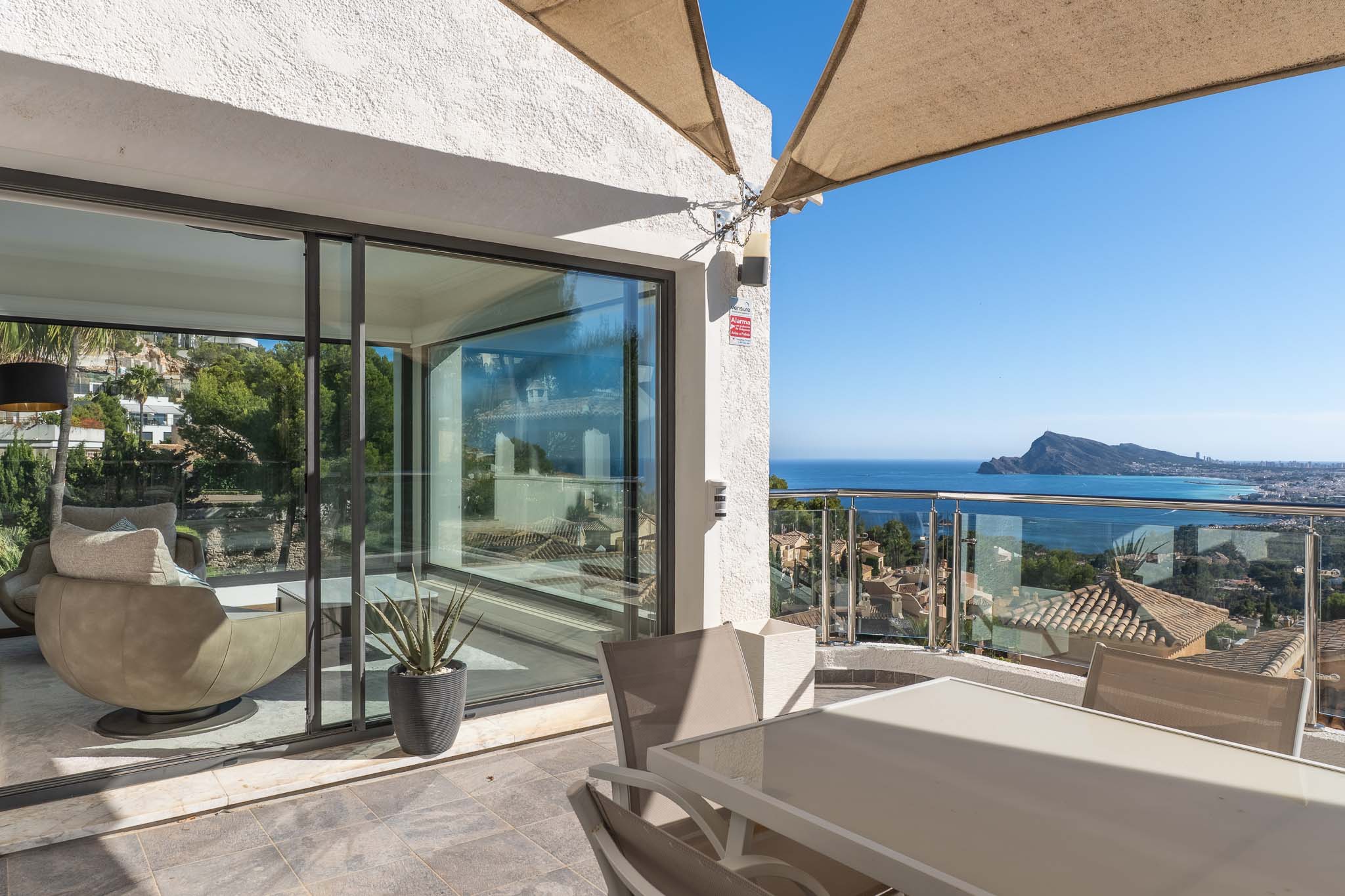 Außenterrasse mit Esstisch und Stühlen, geschützt durch ein Sonnensegel. Bietet Panoramablick auf das Meer und die Küste durch ein Glasgeländer, verbunden mit einem hellen Innenbereich.